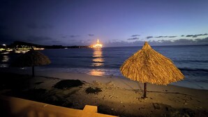 Une plage au crépuscule, avec un parasol en chaume et un bâtiment illuminé au loin.