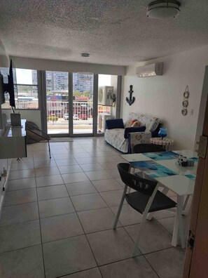 A spacious living area with a large sliding glass door leading to a balcony, a dining table with chairs, and a sofa.