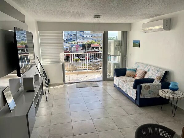 A modern living room with a sofa, a TV, a small table, and a balcony with a view of the city.