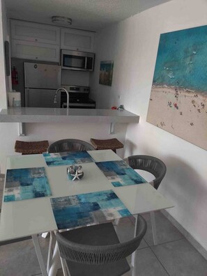 A dining area with a table set for four, a kitchen with modern appliances, and a beach-themed painting on the wall.