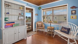 Una habitación con techo de madera, un banco blanco con un cojín que dice 'Beach', una silla de mimbre y un mueble blanco con estantes.