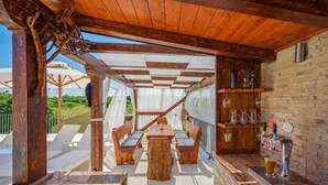 Une terrasse extérieure avec des meubles en bois, un coin bar avec des bouteilles et des verres, et une vue sur la verdure.