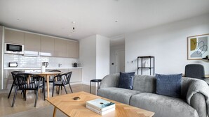 Un salon moderne avec un canapé gris, une table basse en bois et un coin repas doté d’un mur recouvert de carreaux blancs et de chaises noires.