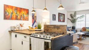 A modern kitchen with a central island, white cabinetry, and a gas stove. There are hanging pendant lights, a potted plant, and artwork on the walls. - Bello Monte Retreat House with Pool in El Yunque (Hato Candal)