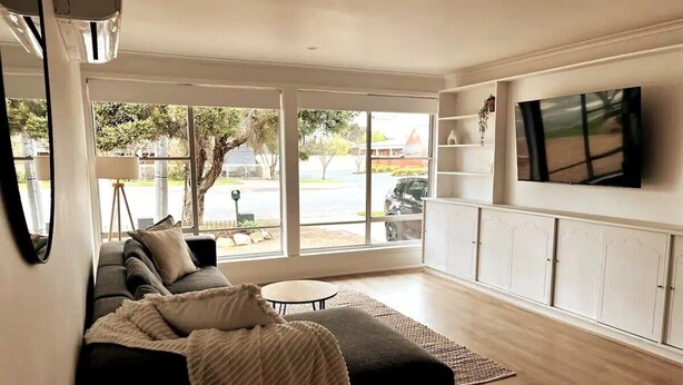 Una sala de estar moderna con un sofá seccional, un televisor montado en la pared y grandes ventanales con vista a la calle.