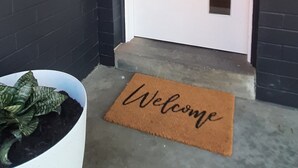 Una puerta oscura con marco blanco, un felpudo con la palabra “Welcome” y una maceta blanca con follaje verde.