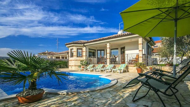 Área da piscina com espreguiçadeiras e guarda-sóis, um pátio de pedra e um edifício com telhado em azulejos.