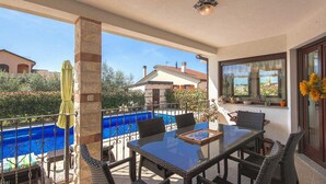 Varanda com mesa de vidro e cadeiras de vime, com vista para uma piscina e uma casa.