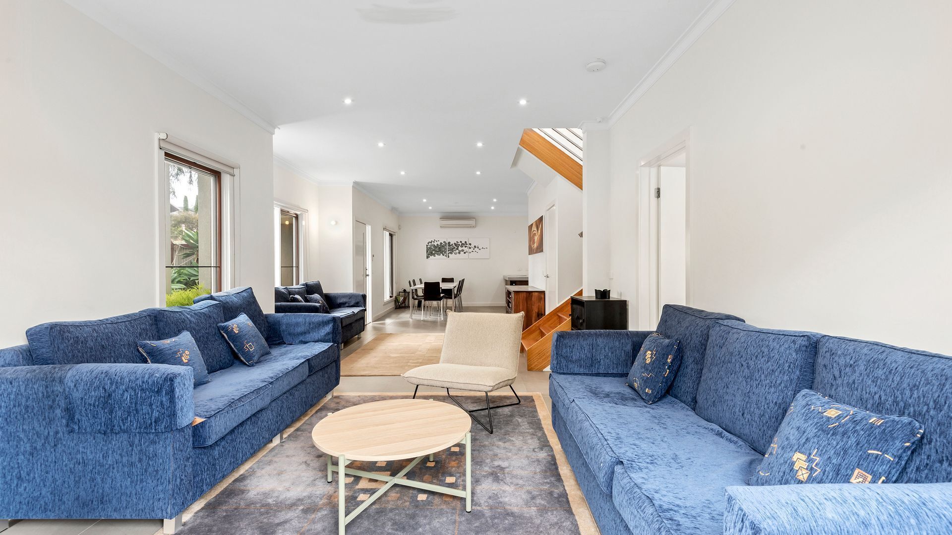 A modern living room with two blue sofas, a small round table, and a dining area in the background.