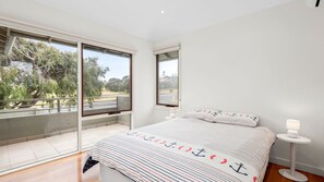 A bedroom with a large window, a bed with striped bedding, a bedside table with a lamp, and a view of trees and a road. - Bayti Retreat - Queenscliff (Queenscliff)