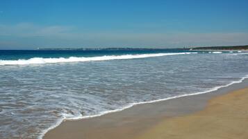 A beach with waves gently lapping onto the shore, a clear sky, and a distant shoreline with buildings.