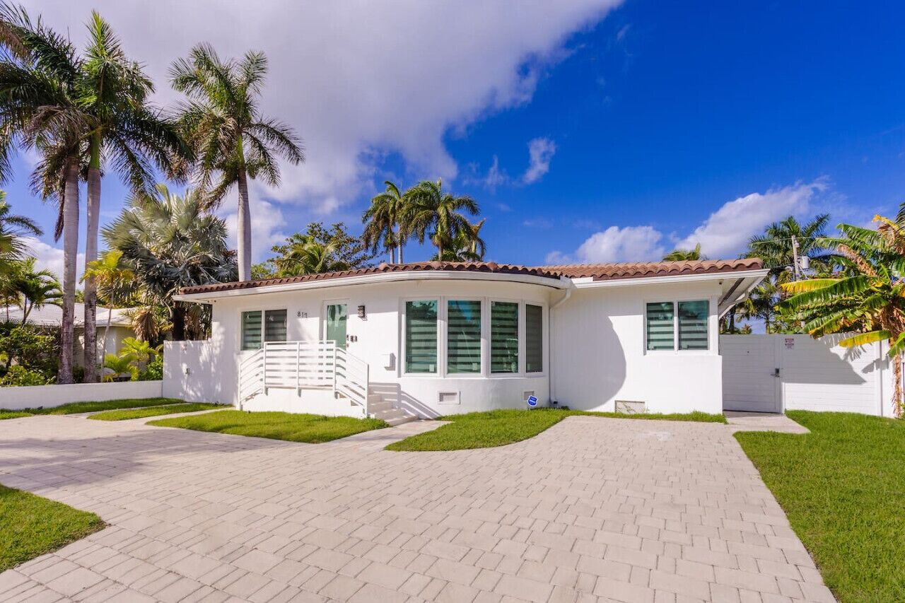 A single-story house with a tiled roof, white exterior walls, and green shutters. It features a front entrance with a set of stairs leading up to a door, a small front yard with grass and a few palm trees, and a paved driveway.