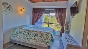 Une chambre à coucher avec un lit, un banc, une télévision et une fenêtre avec des rideaux.