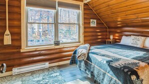 Una habitación de cabaña acogedora con un ventanal grande, una cama con colcha, una mesita con lámpara y un remo sujeto a la pared.