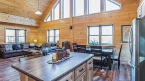 Amplia sala de estar con techo de madera, ventanas grandes, una isla central con pileta y una mesa de comedor con sillas.