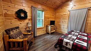 Ein gemütliches Blockhauszimmer mit Holzwänden, einem Bett mit Quilzdecke, einem Sessel mit gemustertem Kissen, einem kleinen Tisch mit Lampe und einem Fernseher auf einem Holzschrank.