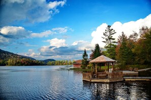 Un pabellón de madera sobre un lago con un muelle, rodeado de árboles y una montaña al fondo.