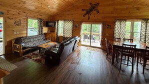 Ein Interieur einer Hütte mit Holzbalkendecke, einer Couch, einem Esstisch und einem an der Wand befestigten Fernseher.