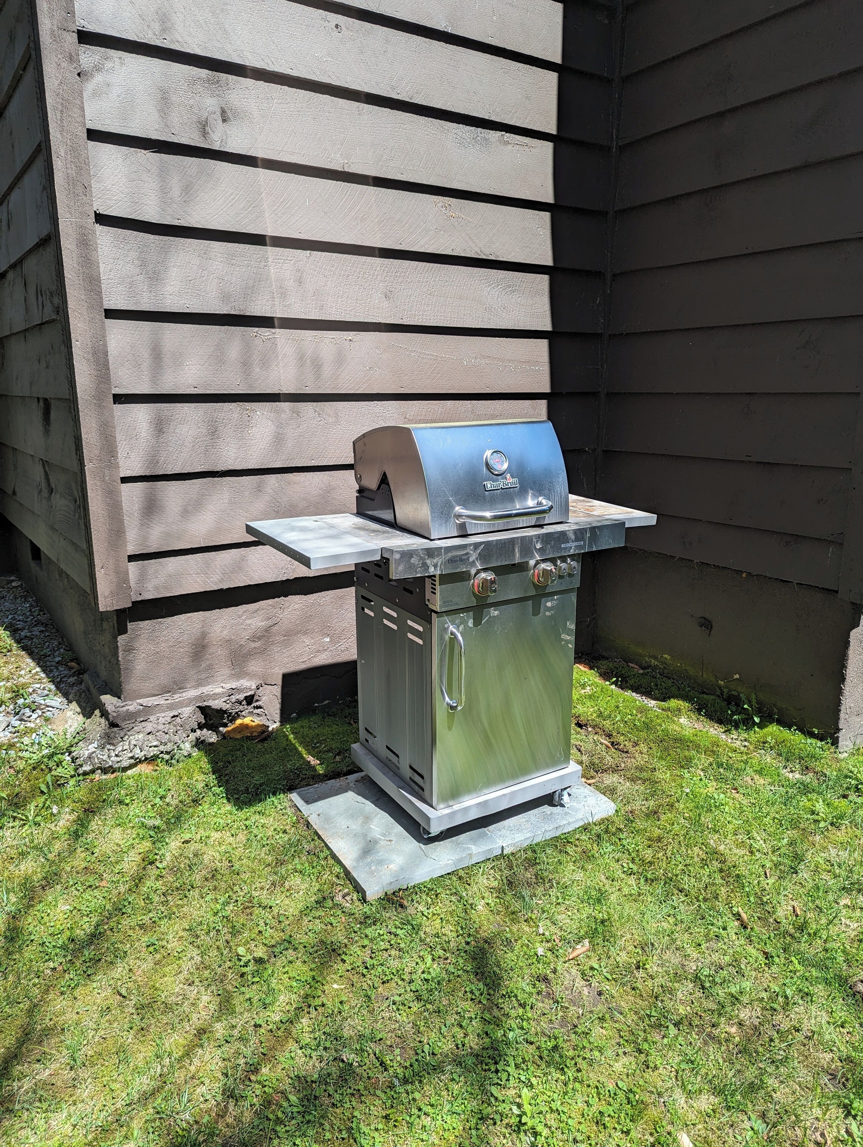A portable stainless steel grill with a digital thermometer, placed on a concrete pad in a grassy area.