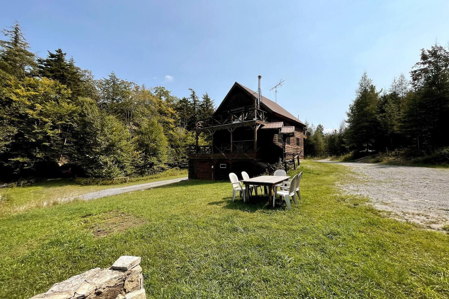 Une cabane en bois rustique, dotée d’une cheminée, entourée d’une zone herbeuse et d’une forêt dense.