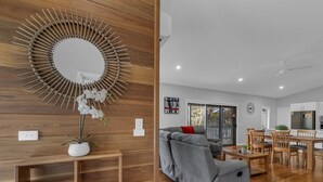 A modern living room with a wooden floor, a grey sofa, a dining area with wooden chairs, and a kitchen in the background.