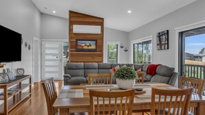 A modern living room with a grey sofa, wooden dining table, and chairs. There is a flat-screen TV mounted on the wall, a potted plant on the table, and a painting above the sofa.