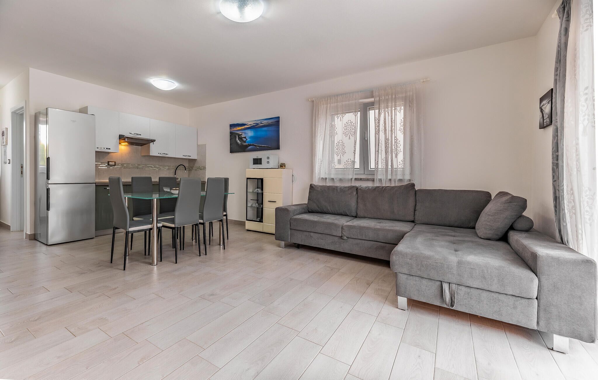 Una sala de estar moderna con un sofá gris, una mesa de comedor con sillas, una zona de cocina con refrigerador y un televisor en la pared.