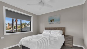 Un dormitorio con un ventanal grande, un ventilador de techo, una cama con ropa blanca, una mesita de noche y un cuadro en la pared.