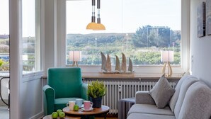 A living room with a teal armchair, a grey sofa, a small round table with cups and apples, and a view of greenery through large windows. - Wohnung Stella - Am Strand- Wohnung Stella (Westerland)