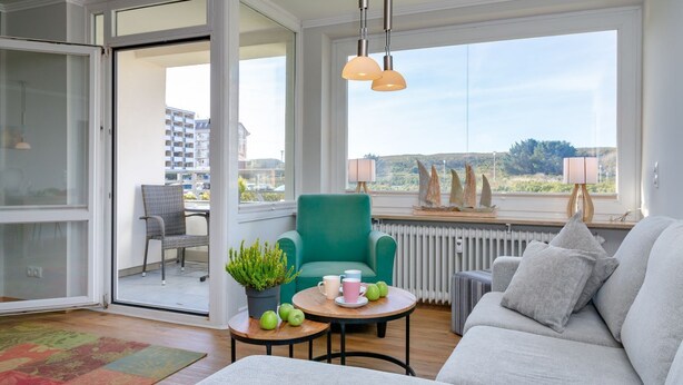 A modern living room with a sofa, a teal armchair, a small table with apples, and a view of a cityscape through large windows. - Wohnung Stella - Am Strand- Wohnung Stella (Westerland)
