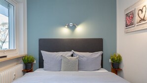 A bedroom with a bed, a headboard, bedside tables with plants, a window, and wall art. - Wohnung Stella - Am Strand- Wohnung Stella (Westerland)