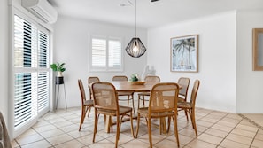 A dining area with a round wooden table surrounded by six wicker chairs, white walls, and a large window with blinds. - Central Location Stylish 3 Bed Pool - Sunshine Central (Sunshine Beach)