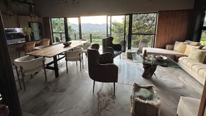 A spacious living area with a dining table, chairs, a sofa, and a coffee table. Large windows offer a view of the outdoors.
