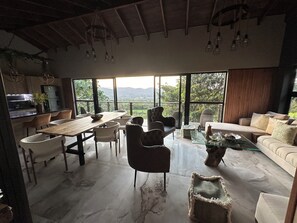 A spacious living area with a dining table, chairs, a sofa, and a coffee table. Large windows offer a view of the outdoors.