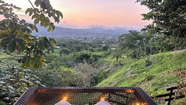 A hammock with two pillows and string lights, set against a backdrop of a lush green landscape and distant mountains.