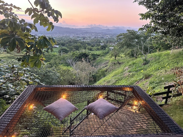 A hammock with two pillows and string lights, set against a backdrop of a lush green landscape and distant mountains.