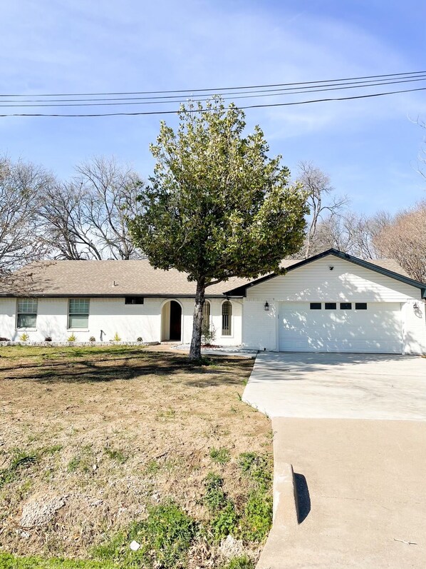 A single-story house with a garage, a tree in front, and a clear sky. - Dallas Fort Worth Getaway Home (Grand Prairie)