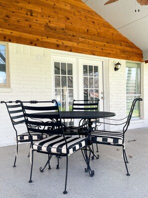 A covered patio with a wooden ceiling, a white brick wall, and a black metal table and chairs. - Dallas Fort Worth Getaway Home (Grand Prairie)