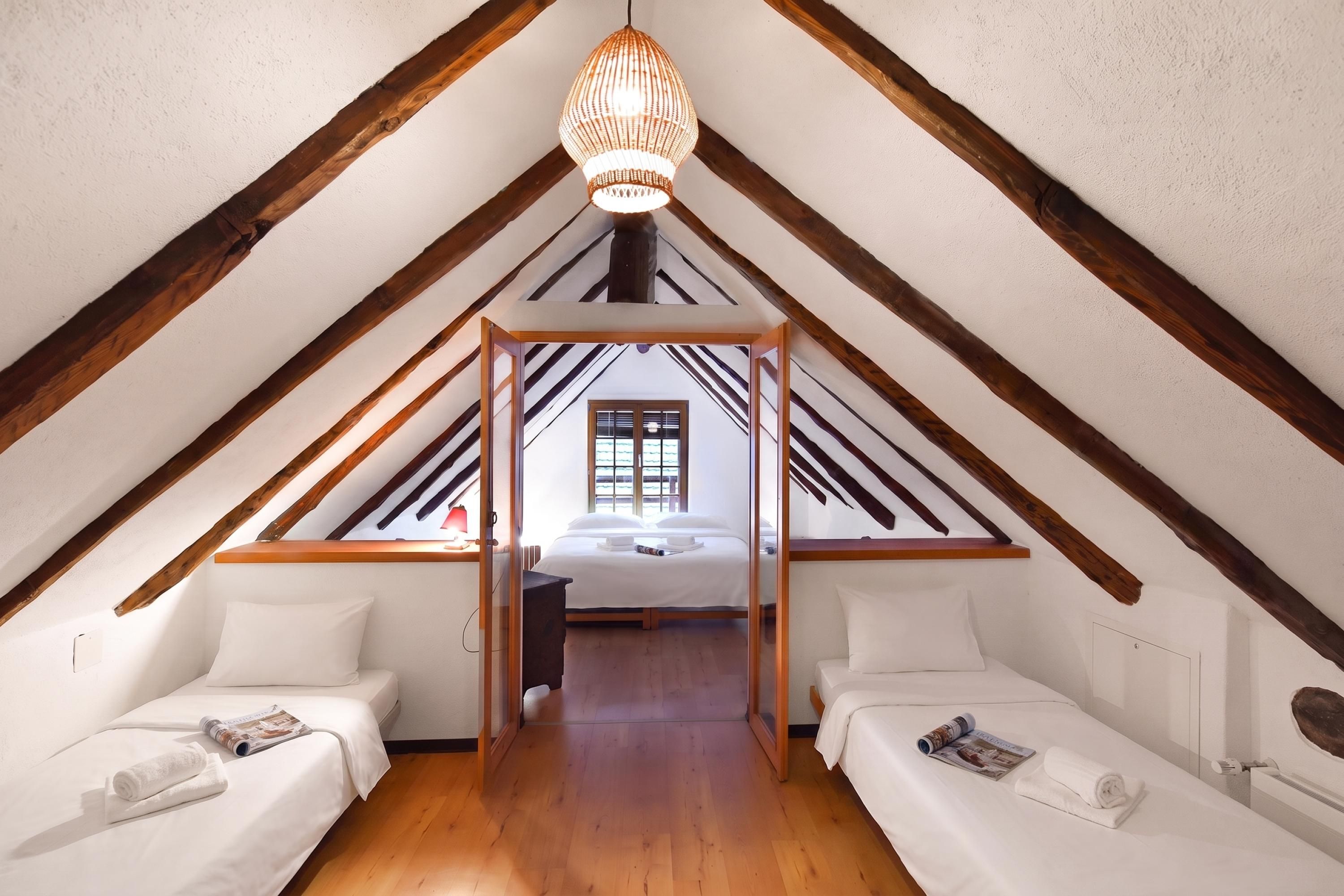 A room with a sloped ceiling, wooden beams, two beds, a hanging light fixture, and a window with blinds.