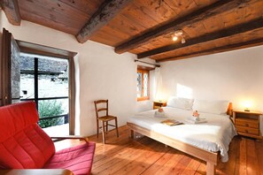 A bedroom with a wooden ceiling, a bed with white bedding, a red chair, a wooden nightstand, and a window with a view of greenery.