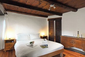 Una cama bien hecha con edredón blanco, una mesita de noche con lámpara, un aparador de madera con una botella y vasos, y un techo de madera.