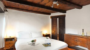 Ein Schlafzimmer mit Holzboden, einem Bett mit weißen Leinentüchern, einem Nachttisch mit Lampe, einer Kommode mit Weingläsern und einer hölzernen Bank.
