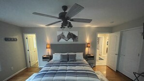 Un dormitorio con una cama, dos mesitas de noche, un ventilador de techo y una puerta que da a otra habitación.