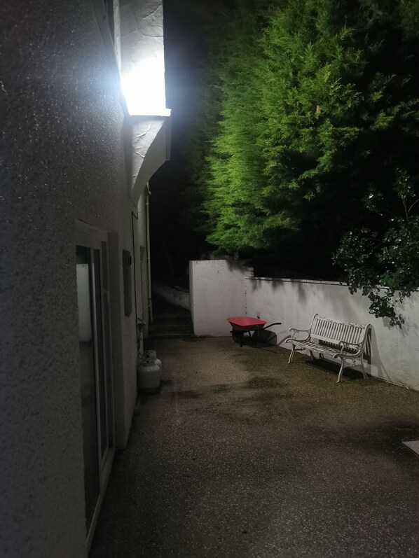 A narrow outdoor corridor with a bench and a wheelbarrow, a wall with a tree, and a bright light source. - The spectacular harbour view (Dunedin)