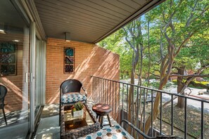 A balcony with a wicker chair, a small table with a plant, and a view of trees and a street. - GP54C-1 |G. Prairie Escape |2BD | Pool | Workspace (Grand Prairie)