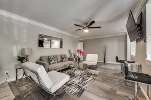 A modern living room with a sofa, a glass coffee table, a white armchair, a massage chair, a ceiling fan, and a flat-screen TV mounted on the wall. - GP54C - Upgraded 2BD Unit | Patio G. Prairie (Grand Prairie)