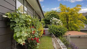Un jardín con diversas plantas y flores, una cerca de madera y una casa con fachada gris.