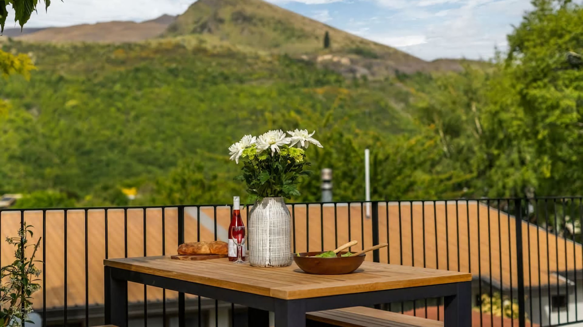 A wooden table with a vase of white flowers, a loaf of bread, a bottle of red wine, and a bowl with a wooden spoon on a balcony with a view of green hills and a mountain.