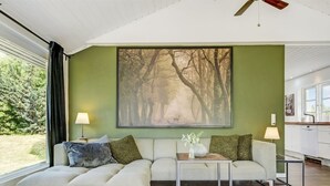 Una sala de estar moderna con un gran ventanal, un sofá, una mesa de centro y un cuadro con una escena de bosque.