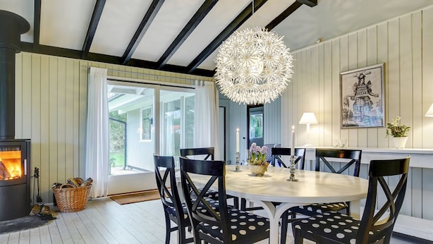 Een eetgedeelte met een ronde tafel, zwarte stoelen met witte stippen, een hanglamp, een open haard en een groot raam met uitzicht op groen.